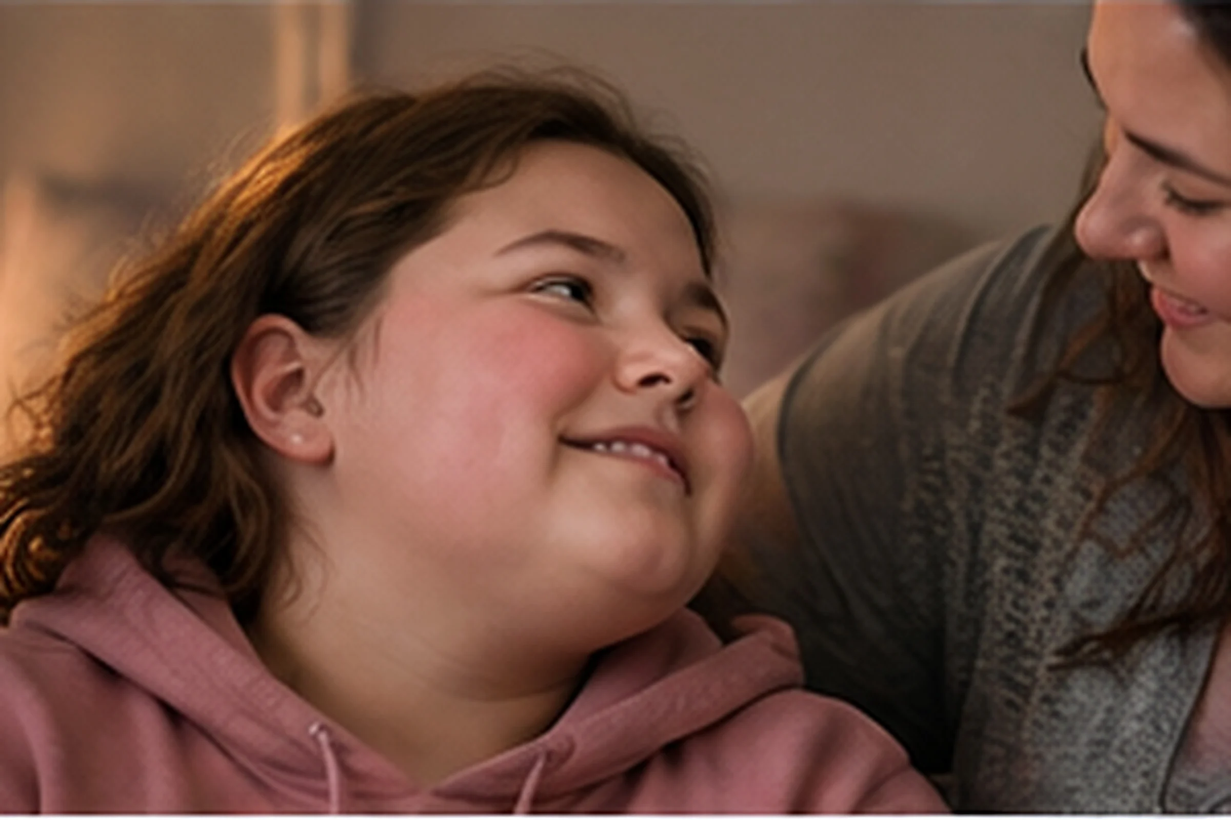 A girl in a pink hoodie smiles up at her mother, who smiles back.