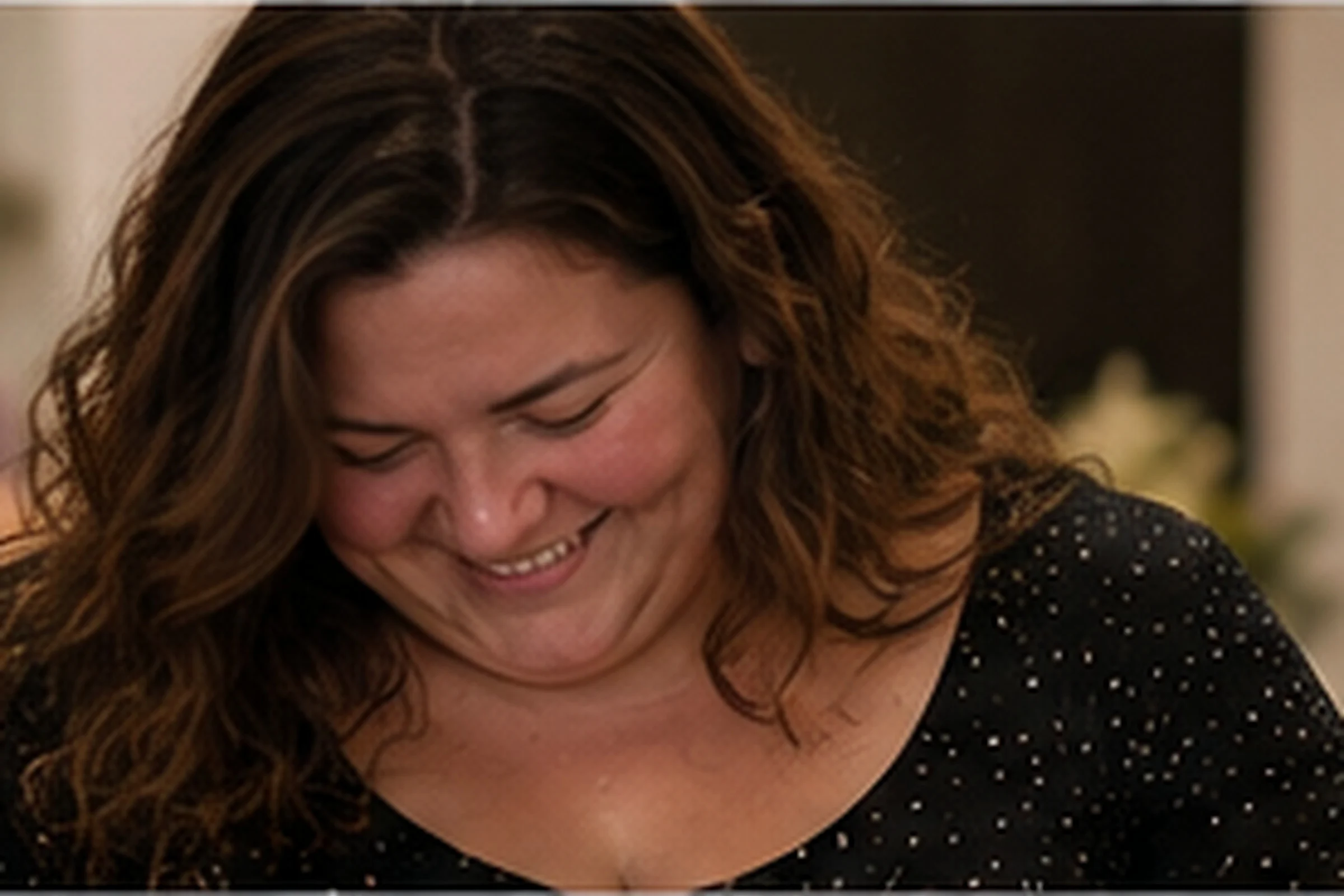 A woman with long dark hair looks down, laughing.