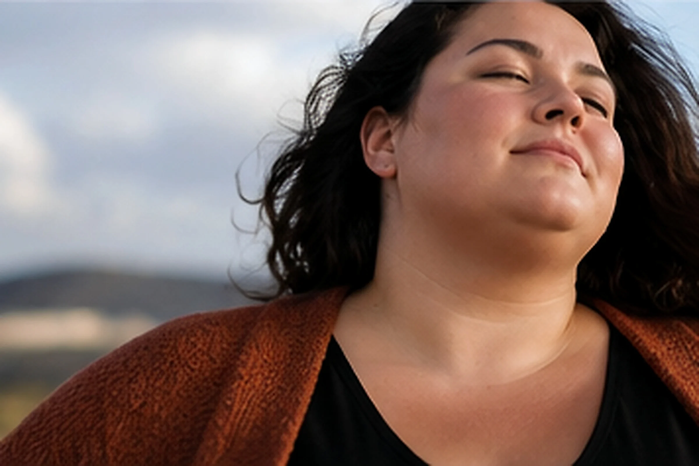 A woman with dark hair and a rust-coloured wrap stands outdoors, eyes closed, face lifted to the sky.
