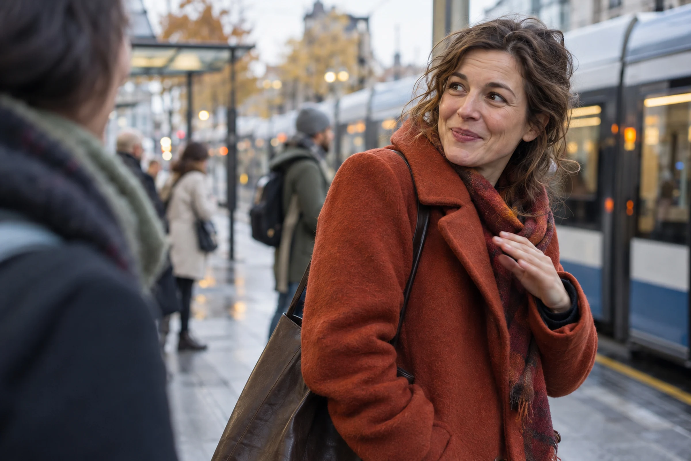 A woman in a rust-coloured coat at a tram stop turns towards someone beside her with a warm, slightly bashful smile, one hand raised to her scarf.