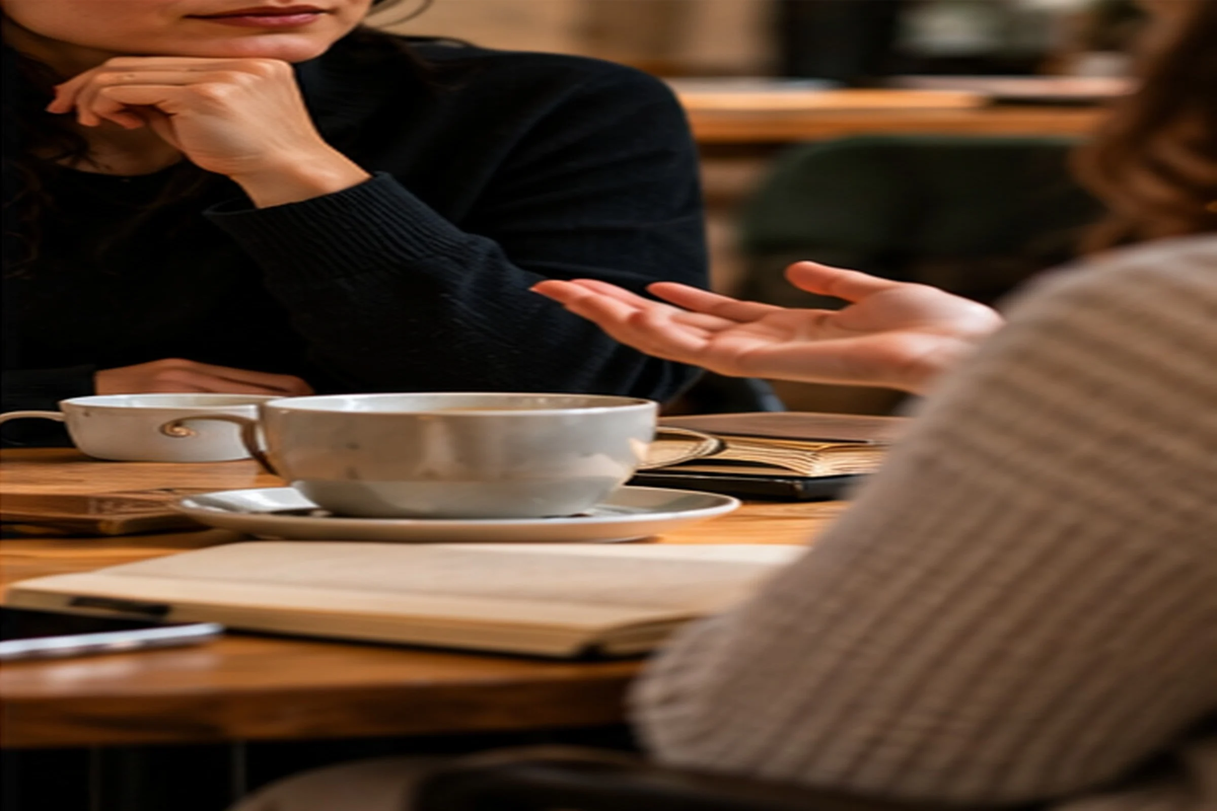 Two people in conversation at a café table with cups of coffee and an open notebook, one resting her chin on her hand while the other gestures as she talks.
